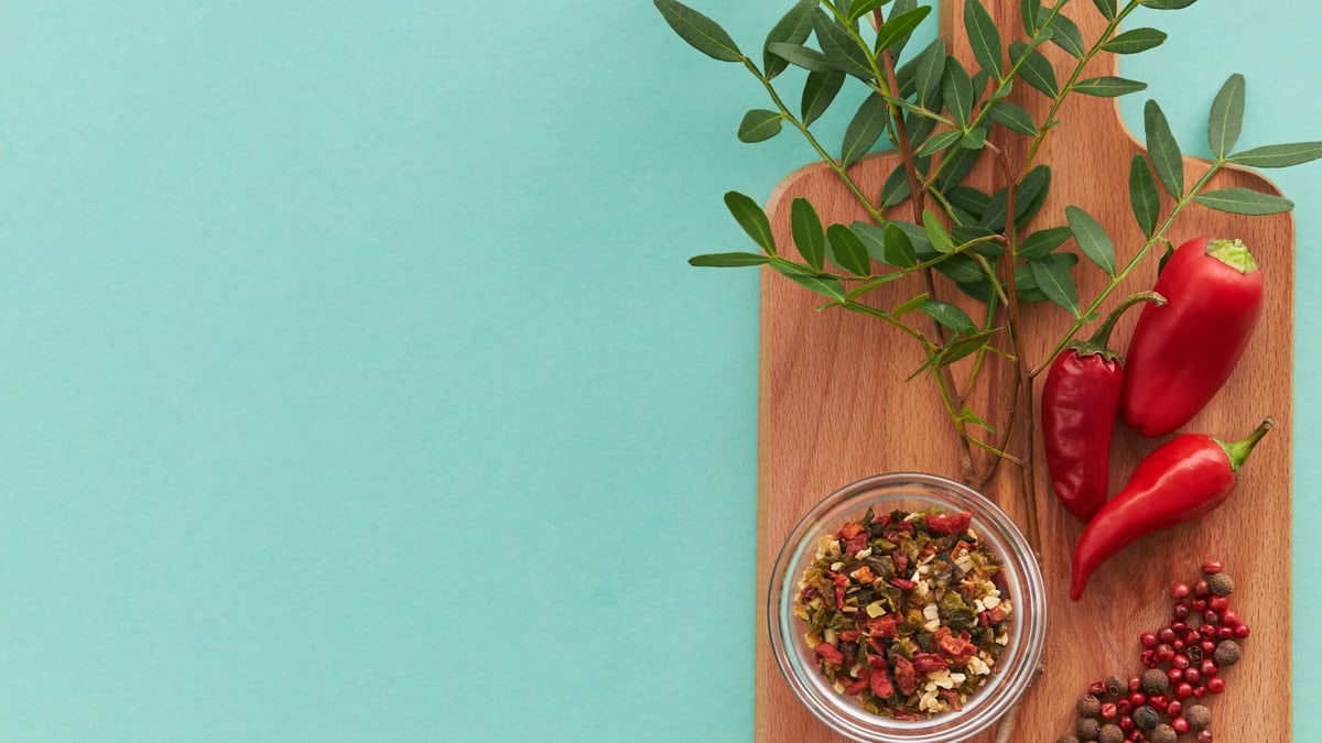 Top view of peppers, spices, and green leaves on a wooden board with teal background, offering copy space.